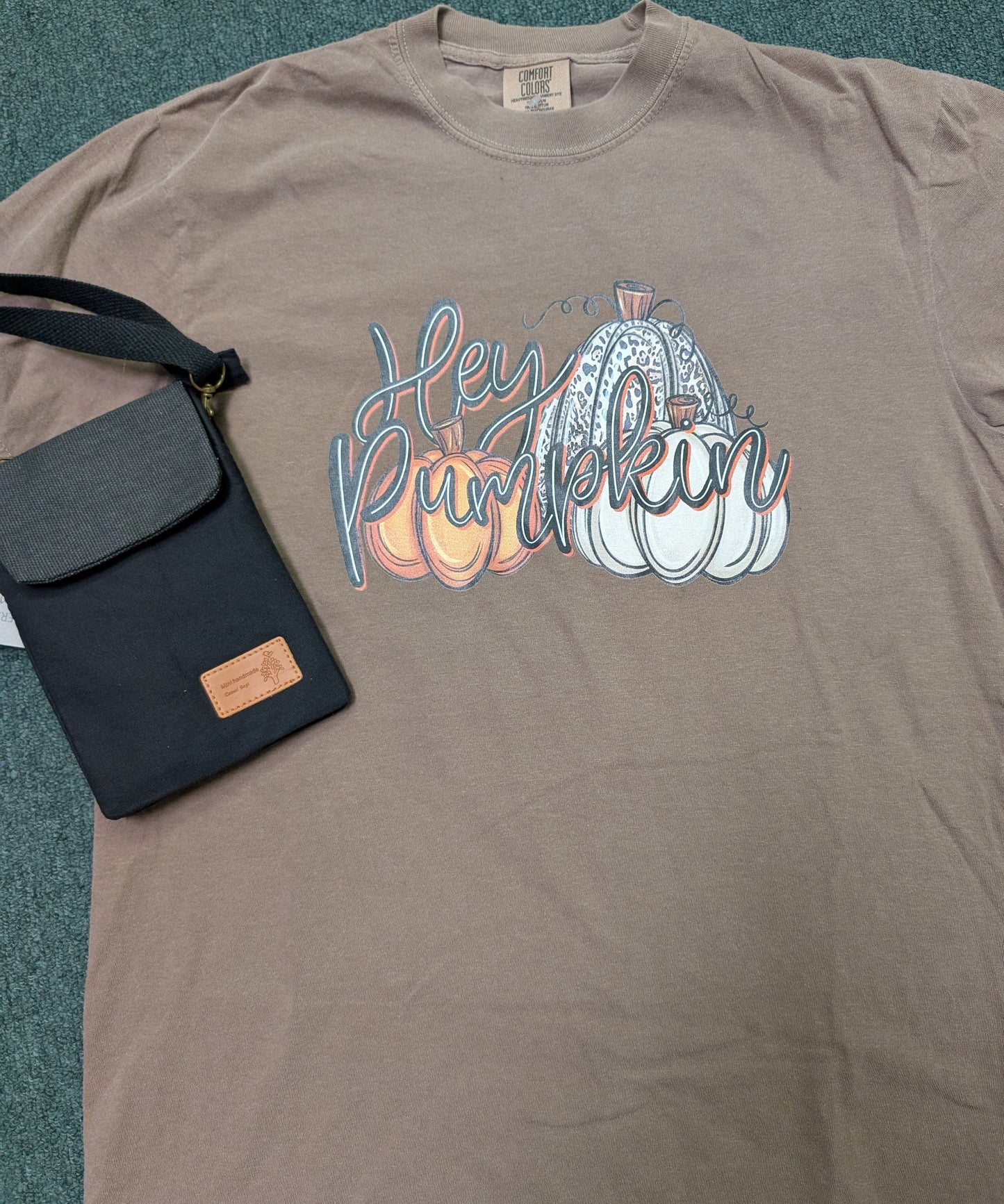 Lt. Brown t-shirt with 'Hey Pumpkin' graphic and black bag on a textured surface